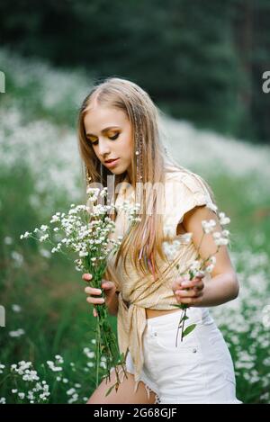 Bella giovane donna che raccoglie fiori bianchi sullo sfondo di un paesaggio forestale in estate. Ritratto di una donna dolce felice in un campo selvaggio, e. Foto Stock