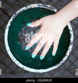 Mano della donna con unghie dipinte blu che toccano l'acqua di una fontana in una città. La fotografia è scattata da un punto di vista sopraelevato ed è in piazza Foto Stock