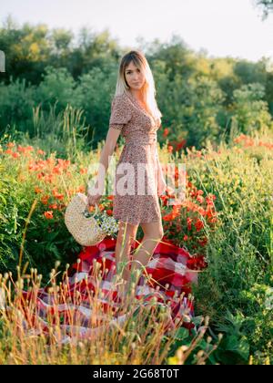 Donna bionda all'aperto con fiori di papavero al tramonto. Donna in abito estivo e borsa in vimini con fiori Foto Stock