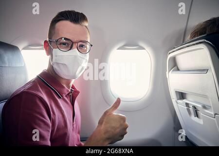 Ritratto di turista felice con maschera facciale durante il volo. Giovane uomo che mostra i pollici in aereo. I temi viaggiano in nuova normale, protezione personale e. Foto Stock
