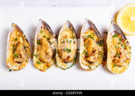 Cozze di greenshell cotte in mezza conchiglia con formaggio, prezzemolo e limone su un piatto bianco. Antipasto caldo tradizionale. Primo piano, vista dall'alto. Foto Stock