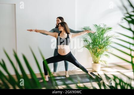 Istruttore di Yoga che aiuta la donna incinta a fare asana guerriero, regolando l'angolo delle braccia. Donna è in piedi su un tappetino in un grande studio, affondo a lato. Lei' Foto Stock