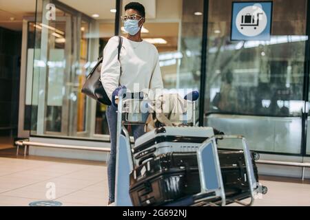 Donna in maschera di faccia che spinge il carrello dei bagagli mentre cammina all'aeroporto. Le donne che viaggiano in pandemia. Foto Stock