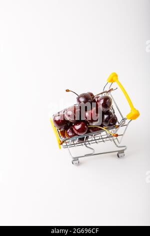 Ciliegia in un piccolo (piccolo) carrello della drogheria per un supermercato. Carrello giocattolo con bacche su sfondo bianco. Primo piano, spazio di copia per il testo, vista dall'alto. Foto Stock