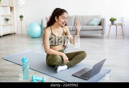 Giovane donna indiana in forma sportiva seduta su tappetino sportivo, salutando il suo personal trainer online su PC portatile, in interni Foto Stock