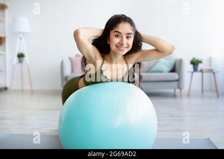 Felice giovane donna indiana che fa esercizi con la palla di fitness, rinforzando i muscoli della schiena a casa Foto Stock