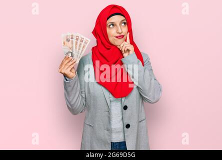 Bella donna ispanica che indossa hijab islamico tenendo 10 libbre banconote faccia seria pensare alla domanda con la mano sul mento, pensieroso circa co Foto Stock