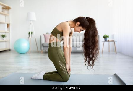 Vista laterale di giovane donna indiana in forma che fa backbend, eseguendo esercizi per la sua salute della spina dorsale a casa, a lunghezza intera Foto Stock