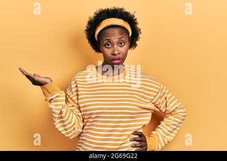 Giovane afroamericana presentandosi con palme aperte, tenendo qualcosa in faccia d'urto, guardando scettico e sarcastico, sorpreso con la bocca aperta Foto Stock