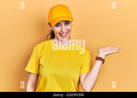 Giovane donna ispanica con uniforme da consegna e cappuccio sorridente allegro presentazione e puntamento con palmo di mano guardando la fotocamera. Foto Stock