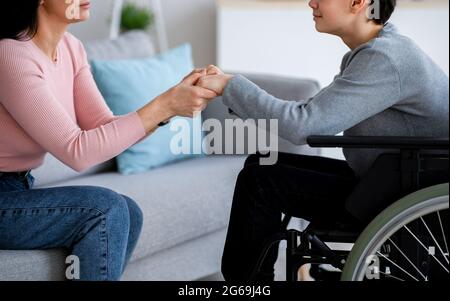 Adolescente portatore di handicap in sedia a rotelle che riceve il supporto dalla madre o da un caregiver, le tiene le mani a casa, closeup Foto Stock