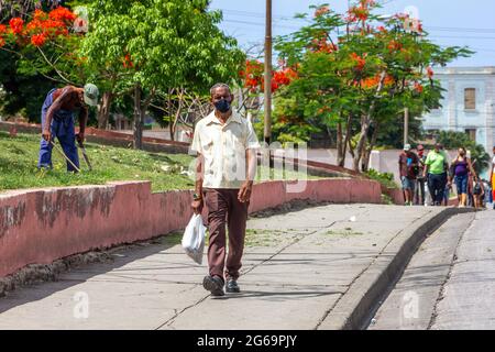 Un uomo cubano che indossa una maschera protettiva cammina sul marciapiede a Santiago de Cuba, Cuba. Un altro uomo lavora tagliando l'erba con un machete nel backgr Foto Stock