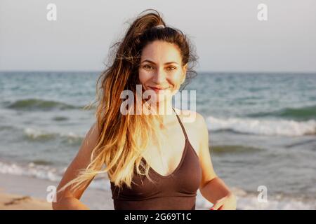 Ritratto di una donna caucasica in forma sportiva che fa il fitness su una spiaggia, indossando reggiseno sportivo, coda di pony, sorridendo alla macchina fotografica. Caldo tramonto estivo sul mare Foto Stock