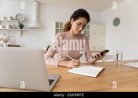 Giovane donna sicura che scrive appunti, utilizzando lo smartphone a casa. Foto Stock