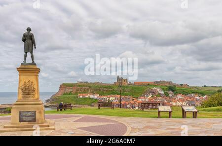 Vista panoramica una scogliera con cottage su una collina e le rovine di un'antica abbazia e una chiesa sopra. Una statua del Capitano Cook è in primo piano Foto Stock