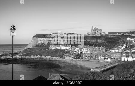 Le case si accoccolano intorno ad un porto con una chiesa e una rovina abbaziale sulla collina sopra. Una spiaggia è sotto e un lampione è in primo piano. Foto Stock