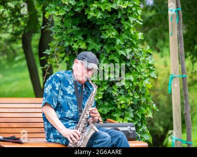 Bucarest, Romania - 06.04.2021: Musicista di strada anziano seduto su una panchina nel parco e suonando al sassofono Foto Stock