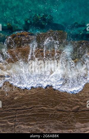 Onde che si infrangono su rocce marroni con marea ascendente, prospettiva drone direttamente sopra Foto Stock