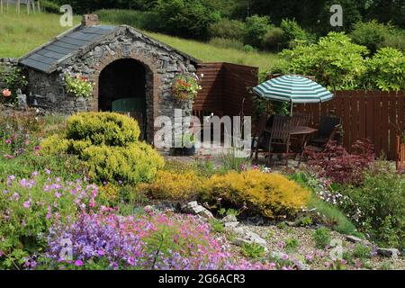 Giardino in Irlanda in estate caratterizzato da rockery in primo piano con fiori in fiore, piccolo edificio in pietra a forge e patio con area salotto Foto Stock