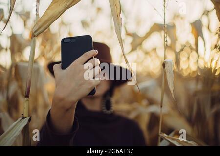 Donna elegante in cappello che prende selfie foto sullo sfondo del campo di mais autunno al tramonto luce del sole, mano con il telefono primo piano. Moda femminile fare s Foto Stock