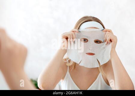 Donna applica il foglio di maschera sul viso. Maschera facciale in tessuto per la cura della pelle. La ragazza teenage fa la terapia di cura della faccia di bellezza. Home trattamenti di bellezza. Igiene quotidiana Self Foto Stock