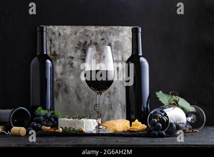 Bicchiere di vino rosso con spuntini e formaggio su sfondo scuro. Bicchiere e bottiglie di vino rosso d'annata su fondo nero in cemento. Foto Stock