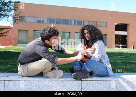 Giovane latino che insegna a suonare la chitarra ad una giovane donna latina nel campus universitario. Vita universitaria, generazione millenaria. Foto Stock