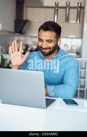 Un giovane indiano positivo in cuffia wireless che si sta muovendo la mano per salutare con una videochiamata Foto Stock