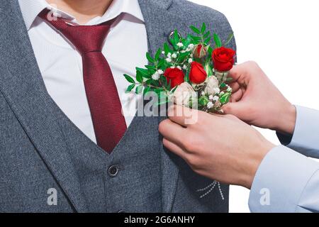 Le mani del ragazzo del testimone al matrimonio attaccano il boutonniere al primo piano del vestito dello sposo. Foto Stock