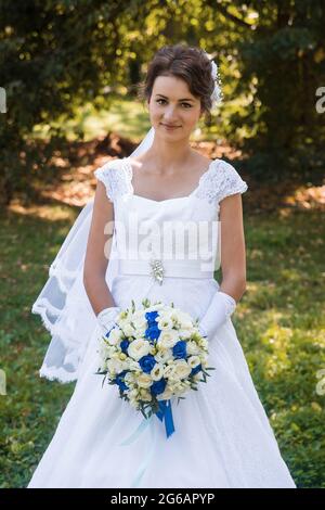 Bielorussia, regione di Minsk - 11 agosto 2018: Bruna giovane e carina sposa di aspetto europeo in un abito da sposa bianco tiene un bouquet di luce e blu f Foto Stock