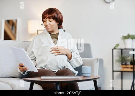 Ritratto minimo di donna afroamericana incinta che legge documenti mentre lavora da casa, spazio di copia Foto Stock
