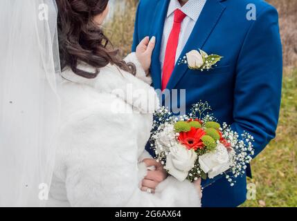 La sposa in un velo in un cappotto bianco si trova di fronte allo sposo in un vestito blu, cravatta rossa e camicia bianca con un mazzo di fiori di nozze. Decorazione esso Foto Stock