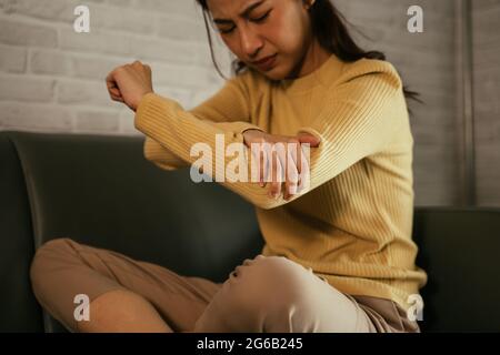Giovane donna asiatica di disagio che tiene la mano e punti di pressione del gomito mentre soffre di dolore articolare del gomito seduto sul divano a casa Foto Stock