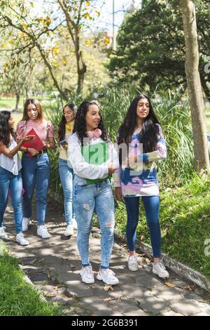 Foto verticale con messa a fuoco selettiva di due amici adolescenti di Latina che camminano verso la scuola superiore con tre fidanzate fuori fuoco dietro un campus Foto Stock