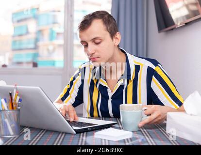 Il giovane si siede di fronte a un computer portatile aperto e beve il tè Foto Stock