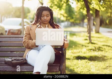 Donna afro-americana freelance seduta su una panchina in una strada e utilizzando un computer portatile Foto Stock