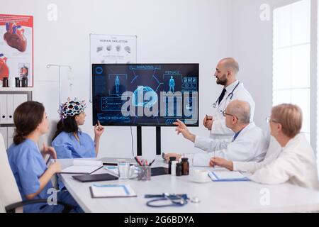 Neurologo uomo medico che controlla l'esperienza cerebrale utilizzando cuffie con sensori su donna assisstant in sala riunioni ospedale. Team medico che analizza il trattamento di malattia esaminando la radiografia medica Foto Stock