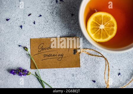 Tazza di tè con una fetta di limone, fiori di lavanda e una buona mattina tag su un tavolo Foto Stock