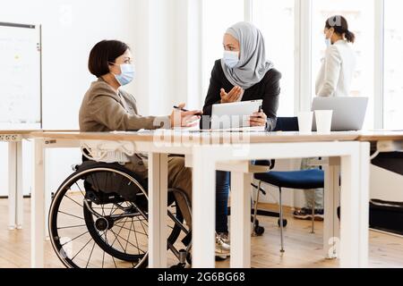 Tre donne in maschera di faccia discutono di progetto mentre lavorano in ufficio Foto Stock