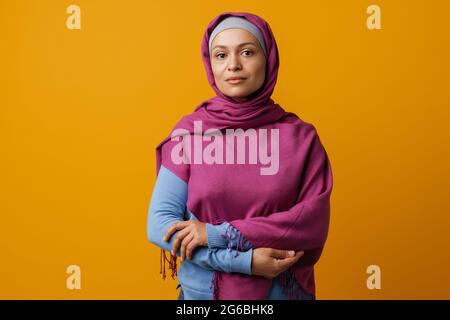 Donna mediorientale in foulard che posa e guarda la telecamera isolata su sfondo giallo Foto Stock