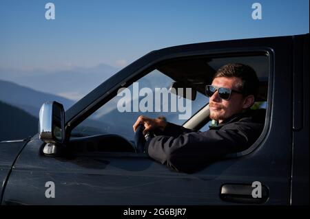 Uomo felice guida SUV nero con occhiali da sole che proteggono il viso dalla luce solare intensa in una calda giornata senza nuvole. Colline di montagna sullo sfondo. Foto Stock