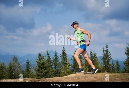 Vista laterale di un ragazzo che corre all'aria aperta da sentieri di montagna. Sentiero estivo che corre maratona in montagna sullo sfondo di alberi verdi, cielo nuvoloso e sagome di montagna beskids. Foto Stock