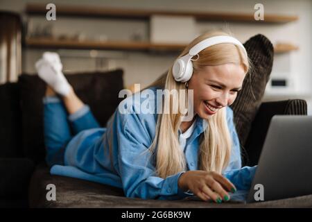 Donna bionda bianca in cuffia che usa il computer portatile mentre si sdraia sul divano a casa Foto Stock