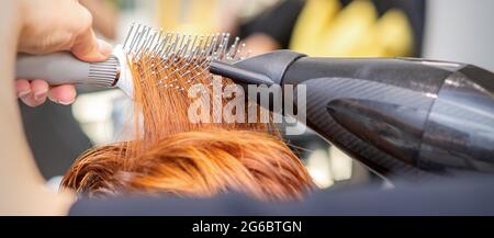 Closeup della mano del padrone con l'aria-asciugando e la spazzola che soffia i capelli rossi femminili in un salone Foto Stock