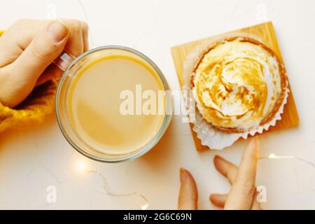 Vista dall'alto della tazza e del tartlet da caffè a contatto con le mani delle donne. Ragazza in maglia maglione sta facendo colazione. Atmosfera romantica. Dolce dessert e bevanda rinfrescante su tavola bianca. Estetica della vita still. Foto Stock