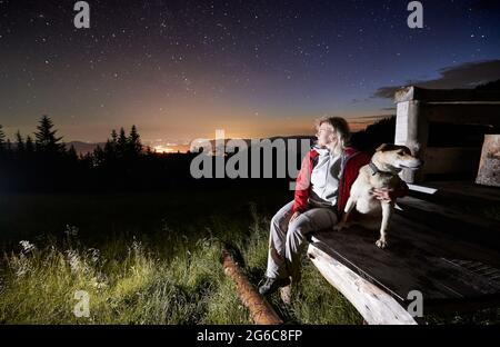 Giovane donna in montagna che guarda bella notte stellata da un portico di  capanna di legno insieme al suo cane. Le luci della città sono  all'orizzonte. Spazio di copia. Concetto di rifugio