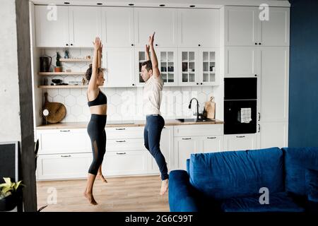 Coppia millennial sportiva concentrata che si diverte in cucina a casa insieme, giovane uomo e donna eccitato attivo che fa sport e salta alzando le mani durante Foto Stock