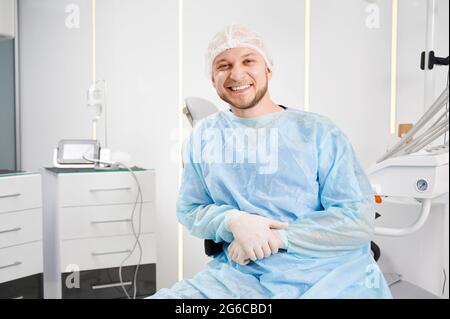 Ritratto di giovane allegro dentista maschile indossando berretto medico, cappotto, guanti seduti nella sua sedia nel suo ufficio moderno. Guardando e sorridendo la fotocamera. Attrezzature dentali high-tech dietro di lui. Foto Stock