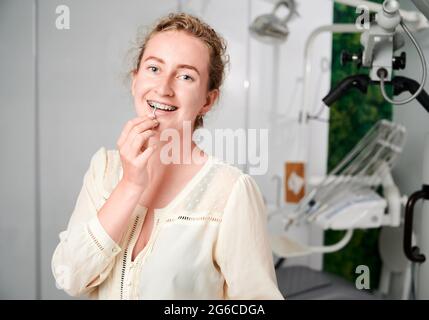 Ritratto di una bella donna in un ufficio dentista ben attrezzato che dimostra spazzolatura interdentale. Giovane ragazza indossa dentali in ceramica e bretelle metalliche. Concetto di odontoiatria, igiene e cura orale. Foto Stock