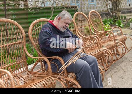 L'uomo ucraino anziano tagliò la manica mentre fa la sedia basculante di vimini nel giardino estivo Foto Stock